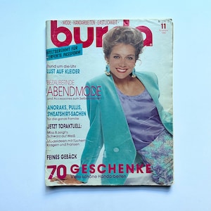 Op de afbeelding: Een vintage Burda magazine cover uit november 1990. De cover toont een vrouw in een teal blazer, een paarse top en een broek met patroon. De titel van het tijdschrift is rood, met tekst in het Duits. De cover bevat ook de tekst "70 GESCHENKE".