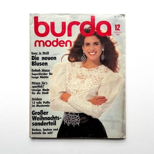 Op de afbeelding: Een vintage Burda Moden magazine cover met een vrouw in een witte blouse met kanten details en een zwarte rok. De titel van het tijdschrift is rood, met Duitse tekst. De cover is van december 1987.