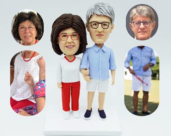 Puede incluir: Un conjunto de figuras personalizadas de una pareja. Las figuras están hechas de arcilla pintada y están de pie sobre una base blanca. La mujer lleva una camisa blanca y pantalones rojos. El hombre lleva una camisa azul y pantalones cortos blancos.