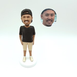 May include: A custom bobblehead figurine of a person wearing a black baseball cap, black t-shirt, khaki shorts, and white sneakers. The figurine has a realistic face and is standing on a white base.