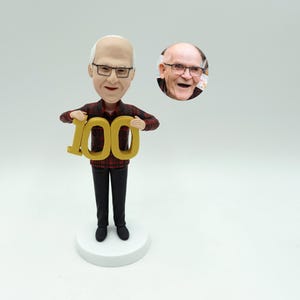 May include: A custom figurine of a person holding a gold number "100". The figurine is wearing a red and black plaid shirt and black pants. The person's face is a photo of an older man with white hair and glasses.