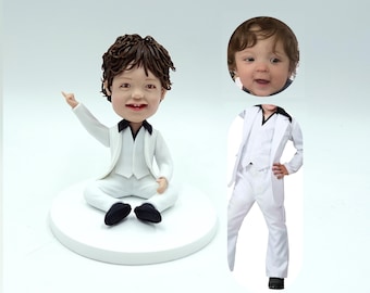 May include: A white ceramic figurine of a child wearing a white suit with a black vest and black shoes. The figurine is sitting with one arm raised and pointing. The child's face is a separate piece and is attached to the figurine. The face is a realistic depiction of a child with brown hair.