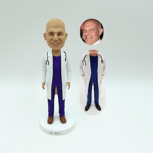 Op de afbeelding: Een gepersonaliseerd beeldje van een dokter die een witte labjas en paarse scrubs draagt. Het beeldje heeft een realistisch gezicht en staat op een witte voet met de naam "Dr. Wine" erop.