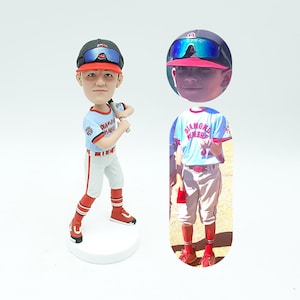 May include: A custom baseball figurine of a young boy wearing a blue and red baseball uniform with the text "Diamond" and "Miners" on the front. The figurine is holding a baseball bat and is standing on a white base. The figurine is based on a photo of the boy in a baseball uniform.