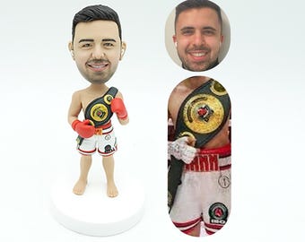 May include: A custom figurine of a boxer wearing a championship belt and red boxing gloves. The figurine is made to look like a real person, with a realistic face and body. The figurine is standing on a white base.