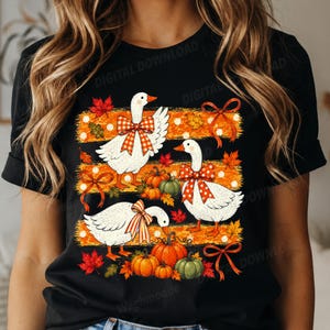 Könnte beinhalten: Ein schwarzes T-Shirt mit einem Grafikdruck von drei weißen Gänsen, die gemusterte Schleifen tragen. Das Design enthält orangefarbene horizontale Streifen, verstreute herbstliche Ahornblätter sowie mehrere orangefarbene und grüne Kürbisse.