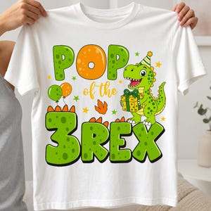 Könnte beinhalten: Ein weißes T-Shirt mit einer bunten Grafik eines grünen Dinosauriers, der einen Partyhut trägt und ein Geschenk hält. Der Text lautet POP of the 3REX, ergänzt durch orangefarbene und grüne Luftballons, kleine Sterne und Fußabdrücke.