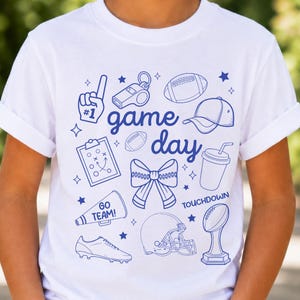 Könnte beinhalten: Ein weißes T-Shirt mit einer blauen Linien-Grafik und dem Text "game day", "GO TEAM!" sowie "TOUCHDOWN". Das Design zeigt Illustrationen von einem Schaumstofffinger, einer Trillerpfeife, Footballs, einer Kappe, einem Klemmbrett, einer Schleife, einem Megafon, einem Schuh, einem Helm und einem Pokal.