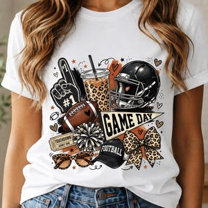 Könnte beinhalten: Ein weißes T-Shirt mit einem Grafikdruck von Football-Motiven, darunter ein schwarzer Helm, ein brauner Football, ein Nummer-eins-Schaumstofffinger, ein Becher mit Leopardenmuster, ein Wimpel mit der Aufschrift GAME DAY, ein Ticket, ein Pompon, eine Kappe und eine Schleife.