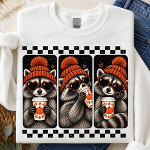 Könnte beinhalten: Ein weißes Sweatshirt mit Rundhalsausschnitt und einem Grafikdruck von drei Waschbären, die orangefarbene Strickmützen und herzförmige Sonnenbrillen tragen. Jeder Waschbär hält einen mit einer Kürbisillustration verzierten Kaffeebecher in den Pfoten.