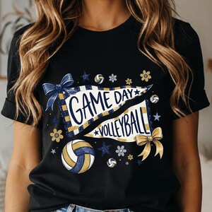 Könnte beinhalten: Ein schwarzes Kurzarm-T-Shirt mit einem Grafikdruck von zwei weißen Wimpeln mit blauen und goldenen Streifen. Der Text lautet "GAME DAY" und "VOLLEYBALL". Das Design enthält blaue und goldene Schleifen, Volleybälle, Sterne und kleine Blumen.