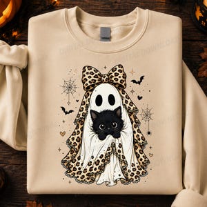May include: A beige crewneck sweatshirt featuring a graphic of a black kitten wearing a white ghost sheet with a leopard print bow and trim. The design includes small black bats, spider webs, and stars surrounding the central ghost kitten figure.