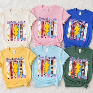Könnte beinhalten: Sechs kurzärmelige T-Shirts in Creme, Pink, Blau, Gelb, Hellblau und Grün. Jedes Shirt zeigt eine Grafik mit Stiften, einem Blitz und dem Text Kindergarten, Second grade, Pre-K, First grade, Fourth grade und Third grade.