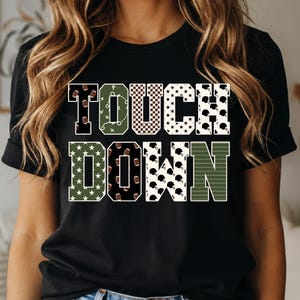 Könnte beinhalten: Ein schwarzes Kurzarm-T-Shirt mit dem Schriftzug TOUCH DOWN in großen Blockbuchstaben. Jeder Buchstabe hat ein einzigartiges Muster mit Footballs, Spielzügen, Sternen, Karos und Footballhelmen in den Farben Grün, Schwarz, Weiß und Braun.