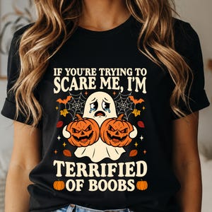 Könnte beinhalten: Ein schwarzes T-Shirt mit einer Grafik eines weinenden weißen Geistes, der zwei orangefarbene Kürbisse hält, umgeben von Spinnweben, Fledermäusen und Blättern. Der Text lautet: IF YOU'RE TRYING TO SCARE ME, I'M TERRIFIED OF BOOBS.