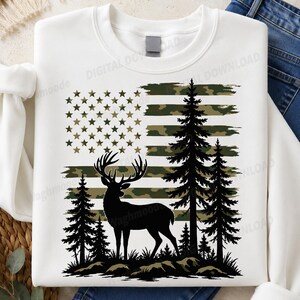 Könnte beinhalten: Ein weißes Sweatshirt mit Rundhalsausschnitt und einem Grafikdruck, der die schwarze Silhouette eines Hirsches vor drei Tannen zeigt. Hinter dem Hirsch und den Bäumen befindet sich ein stilisiertes US-Flaggenmuster in grünen Camouflage-Tönen.