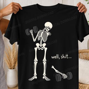 Könnte beinhalten: Ein schwarzes T-Shirt mit einer weißen Grafik eines Skeletts, das Hanteln hebt. Ein Arm des Skeletts ist an der Schulter abgetrennt, wobei die Hand noch eine Hantel auf dem Boden hält. Der Text "well, shit..." ist in Weiß gedruckt.