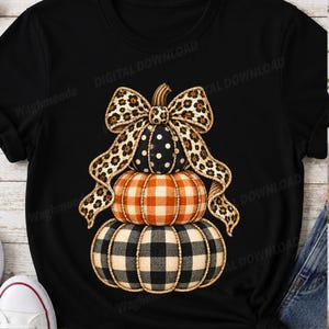 May include: A black t-shirt featuring a graphic of three stacked pumpkins. The top pumpkin is black with white polka dots, the middle is orange plaid, and the bottom is black and white buffalo check. A large leopard print bow sits on top.