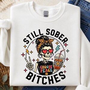 May include: A white sweatshirt featuring a graphic of a skeleton wearing a leopard print headband and heart-shaped sunglasses. The skeleton holds a drink and makes a peace sign. The text reads STILL SOBER BITCHES and CHOOSE PEACE.
