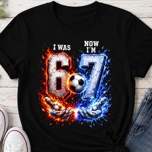 Könnte beinhalten: Ein schwarzes Kurzarm-T-Shirt mit einem Grafikdruck und dem Text I WAS 6 sowie NOW I'M 7. Das Motiv zeigt einen Fußball zwischen zwei Händen, umgeben von leuchtend roten Flammen auf der linken und blauer Energie auf der rechten Seite.