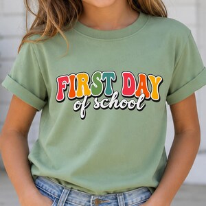 Könnte beinhalten: Ein kurzärmeliges T-Shirt in Salbeigrün mit dem aufgedruckten Text "FIRST DAY of school" auf der Vorderseite. Das Wort "FIRST" ist in großen, bunten Blockbuchstaben im Retro-Stil gehalten, während "of school" darunter in weißer Schreibschrift steht.