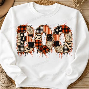Könnte beinhalten: Ein weißes Langarm-Sweatshirt mit dem Wort BOO in einem Patchwork-Design. Die Buchstaben bestehen aus verschiedenen Stoffmustern, darunter orange-schwarzes Karomuster, Spinnennetze, Geister, Fledermäuse und Kürbisse mit ausgefransten Rändern.