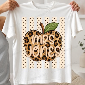Könnte beinhalten: Ein weißes T-Shirt mit kurzen Ärmeln und einem zentralen Apfel-Motiv mit Leopardenmuster. Der Apfel hat ein grünes Blatt und den Schriftzug "MRS JONES" in heller Schrift. Der Hintergrund des Motivs besteht aus vertikalen Streifen.