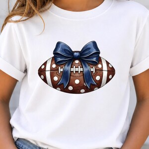 Könnte beinhalten: Ein weißes Kurzarm-T-Shirt mit einer Grafik eines braunen Footballs mit weißen Punkten und weißen Streifen. Eine große, dunkelblaue Schleife ist oben auf dem Football platziert und mittig über den Schnürsenkeln des Balls angeordnet.