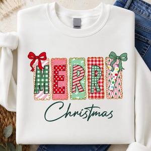 Könnte beinhalten: Ein weißes Sweatshirt mit Rundhalsausschnitt und dem Wort MERRY in großen, blockartigen Buchstaben mit verschiedenen Mustern wie grünem Vichy-Karo, roten Punkten, Zuckerstangenstreifen, rotem Schottenkaro und bunten Bäumen. Rote und grüne Schleifen verzieren den Text.