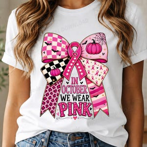 Könnte beinhalten: Ein weißes T-Shirt mit einer großen Grafik einer rosa Schleife. Das Design der Schleife enthält Muster aus rosa Kürbissen, Fledermäusen, Spinnennetzen, Leopardenmuster und Streifen. Der Text lautet IN OCTOBER WE WEAR PINK in rosa und schwarzer Schrift.