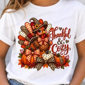 Könnte beinhalten: Ein weißes T-Shirt mit einer Grafik eines Truthahns mit Leopardenmuster-Hut, umgeben von Kürbissen, Herbstblättern und Beeren. Der Text Thankful & Cozy ist in roter Schreibschrift gehalten, ergänzt durch kleine Herzen und Karoschleifen.