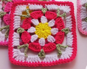 May include: Handmade crochet coasters in a floral design. The coasters feature a square shape with a pink border, white and pink flower details, and a yellow center. A mug with a bee design sits on one of the coasters. The image also includes the text "CROCHET PATTERN ENG PDF".