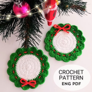 Könnte beinhalten: Zwei grüne und weiße Häkeluntersetzer mit roten Schleifen. Die Untersetzer sind wreath-förmig und haben eine weiße Mitte mit einem grünen Außenring. Der Text "CROCHET PATTERN ENG PDF" ist im Bild sichtbar.