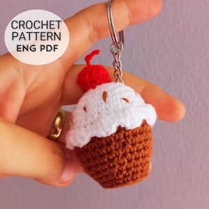 Puede incluir: Un llavero de cupcake tejido a crochet con una cereza roja en la parte superior. El llavero es marrón y blanco y está unido a un llavero plateado.