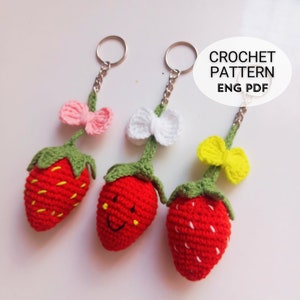 Könnte beinhalten: Drei gehäkelte Erdbeer-Schlüsselanhänger mit verschiedenen farbigen Schleifen. Die Erdbeeren sind rot mit weißen Nähten und grünen Blättern. Die Schleifen sind rosa, weiß und gelb.
