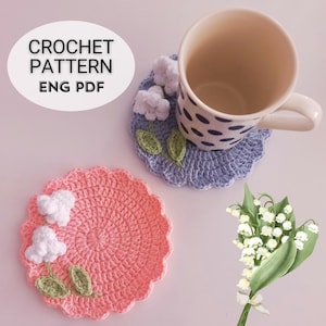 May include: Two crochet coasters, one pink and one blue, with floral embellishments. The pink coaster has white flowers and green leaves. The blue coaster has white flowers and green leaves, with a mug on top. The image includes the text "CROCHET PATTERN ENG PDF".
