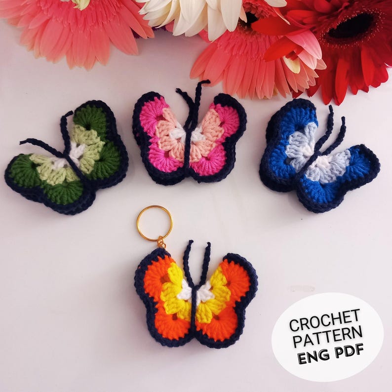 May include: A collection of colorful, crocheted butterfly decorations. The butterflies feature various color combinations, including green, pink, blue, and orange, with dark blue outlines. One butterfly is attached to a gold keychain. The image also includes text that reads "CROCHET PATTERN ENG PDF."
