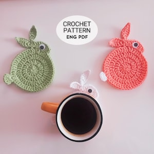 Może przedstawiać: Trzy ręcznie robione podkładki pod kubki w kształcie królika w kolorze zielonym, koralowym i białym, z ruchomymi oczami. Kubek kawy spoczywa na białej podkładce w kształcie królika. Obraz zawiera tekst "CROCHET PATTERN ENG PDF".