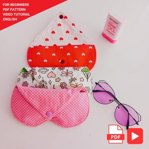 May include: Three fabric eyeglass cases with snap closures. The top case is white with red hearts, the middle case features a floral and dragonfly pattern, and the bottom case is pink with white polka dots. A pair of purple-tinted glasses is included.