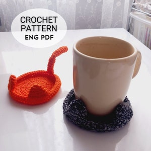 May include: A crocheted orange cat-shaped coaster with a tail and a gray crocheted cat-shaped coaster with two paws. The gray coaster is holding a white mug with coffee inside. The text "CROCHET PATTERN ENG PDF" is written in black on a white oval.