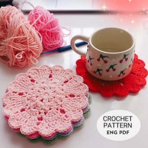 May include: A collection of crochet items, including a stack of pink, purple, and green coasters, a red coaster, and a mug with a cherry pattern. Pink and peach yarn balls and a crochet hook are also present. The image includes the text "CROCHET PATTERN ENG PDF."