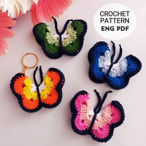 May include: A collection of colorful, crocheted butterfly decorations. One butterfly is attached to a gold keychain. The butterflies are in various colors, including orange, yellow, blue, green, and pink, with dark blue outlines. The text "CROCHET PATTERN ENG PDF" is in a white circle.