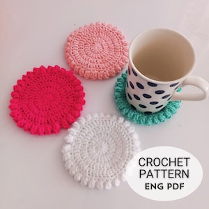 Może przedstawiać: Cztery dzianinowe podkładki pod kubki w kolorze różowym, czerwonym, białym i zielonym. Podkładki mają okrągły kształt z wypukłym środkiem i falistym brzegiem. Biały kubek w czarne kropki stoi na zielonej podkładce.