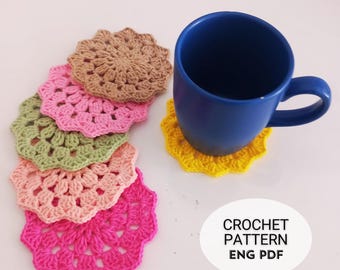 May include: A set of six crocheted coasters in various colors, including pink, green, brown, and yellow. A blue mug is sitting on a yellow coaster. The text "CROCHET PATTERN ENG PDF" is visible in the image.