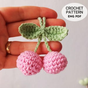 May include: A crocheted cherry pattern with two pink cherries and green leaves. The text "CROCHET PATTERN ENG PDF" is in a white circle. The cherries are held in a hand.