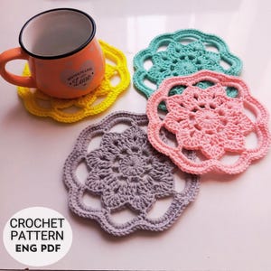 以下が含まれることがあります： 黄色、ティール、ピンク、グレーの4つの手作りのかぎ針編みコースター。花柄のデザインです。ハートと「Homemade with Love」の文字が入ったオレンジ色のマグカップがコースターの上に置かれています。「CROCHET PATTERN ENG PDF」の文字が左下にあります。