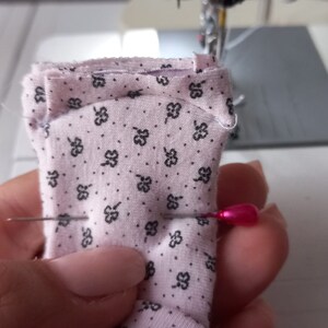 May include: A close-up of a pink fabric with a small black floral pattern being sewn on a sewing machine. A pink pin is holding the fabric in place.