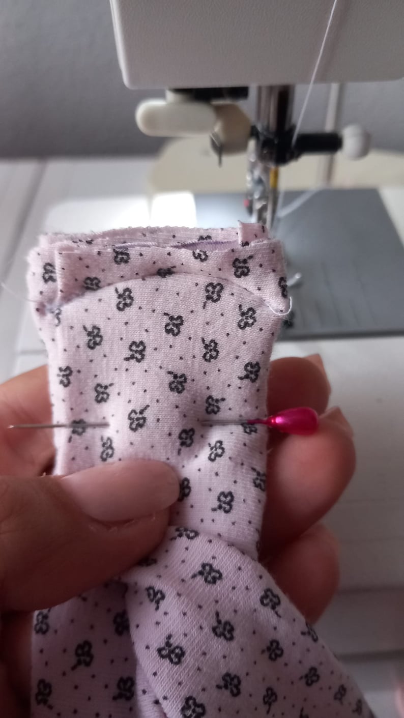 May include: A close-up of a pink fabric with a small black floral pattern being sewn on a sewing machine. A pink pin is holding the fabric in place.