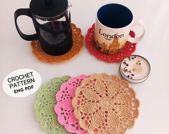 Puede incluir: Cuatro posavasos de ganchillo en verde, rosa, beige y rojo. Los posavasos son redondos y tienen un delicado patrón de encaje. Una cafetera y una taza están encima de los posavasos. La taza tiene la palabra "London" impresa.