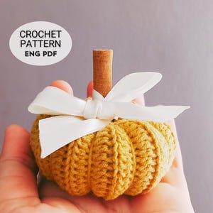 May include: A crocheted pumpkin in shades of yellow-orange, adorned with a white ribbon and a cinnamon stick stem. The pumpkin is handmade, with visible stitch details. The image includes text that reads "CROCHET PATTERN ENG PDF".